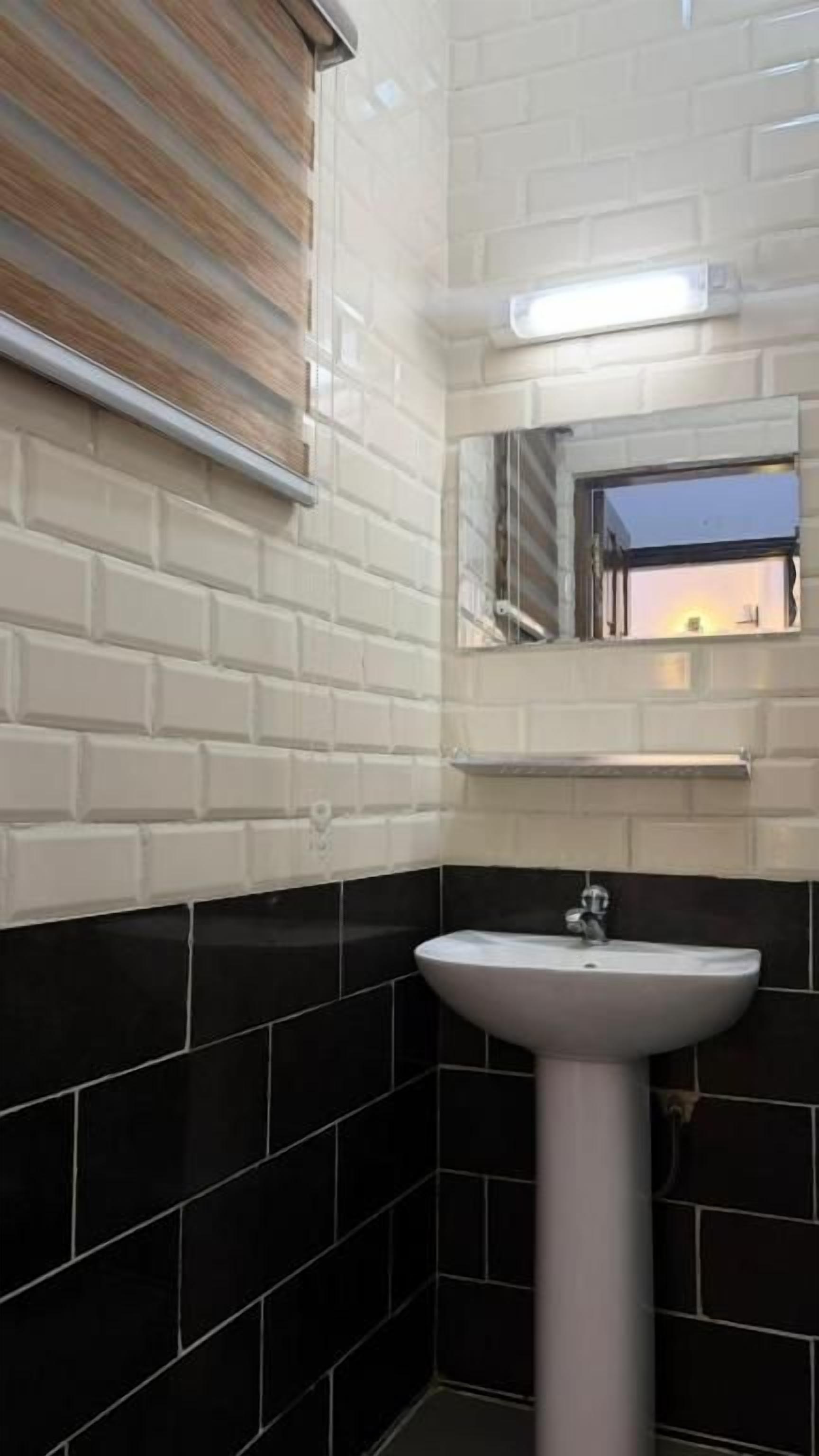 Un baño con un lavamanos pedestal blanco, paredes con azulejos negros, un ventanal con persianas y un espejo sobre el lavamanos.