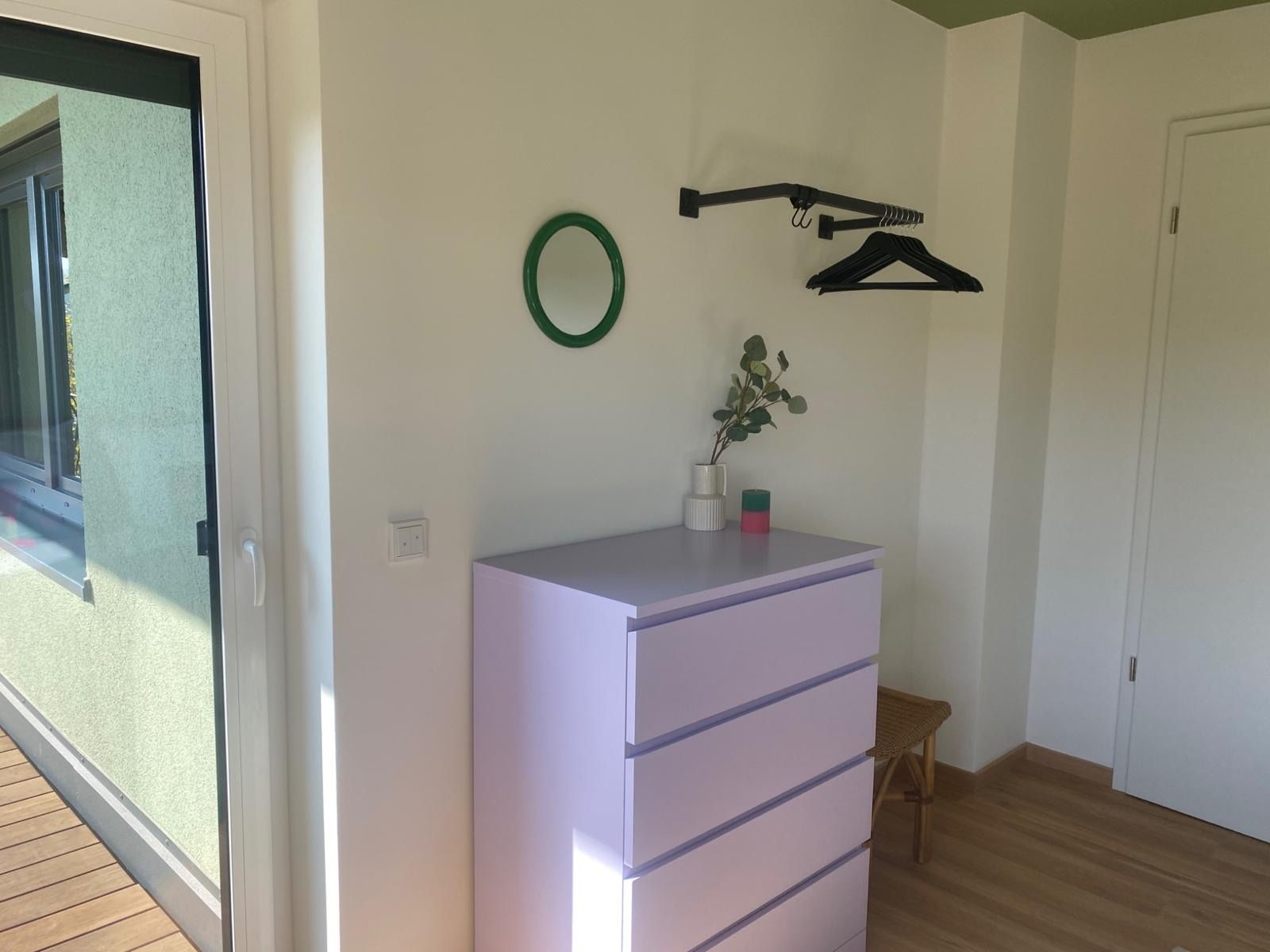 A room with a sliding glass door, a white dresser with drawers, a small wooden stool, and a wall-mounted coat rack with a green circular object.