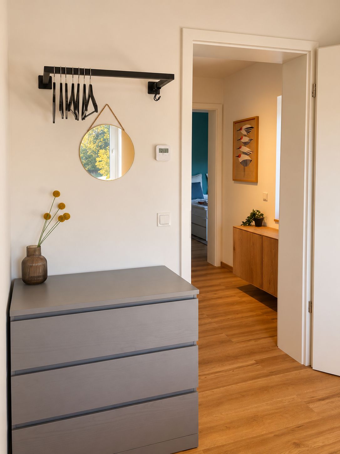 A hallway with a grey dresser, a round mirror, a wall clock, and a door leading to another room.