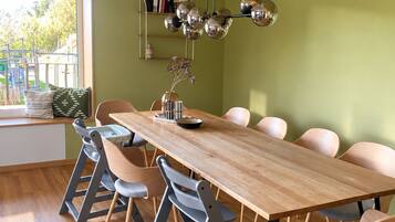 A dining area with a long wooden table, six chairs, a hanging spherical light fixture, and a window with a view of greenery.