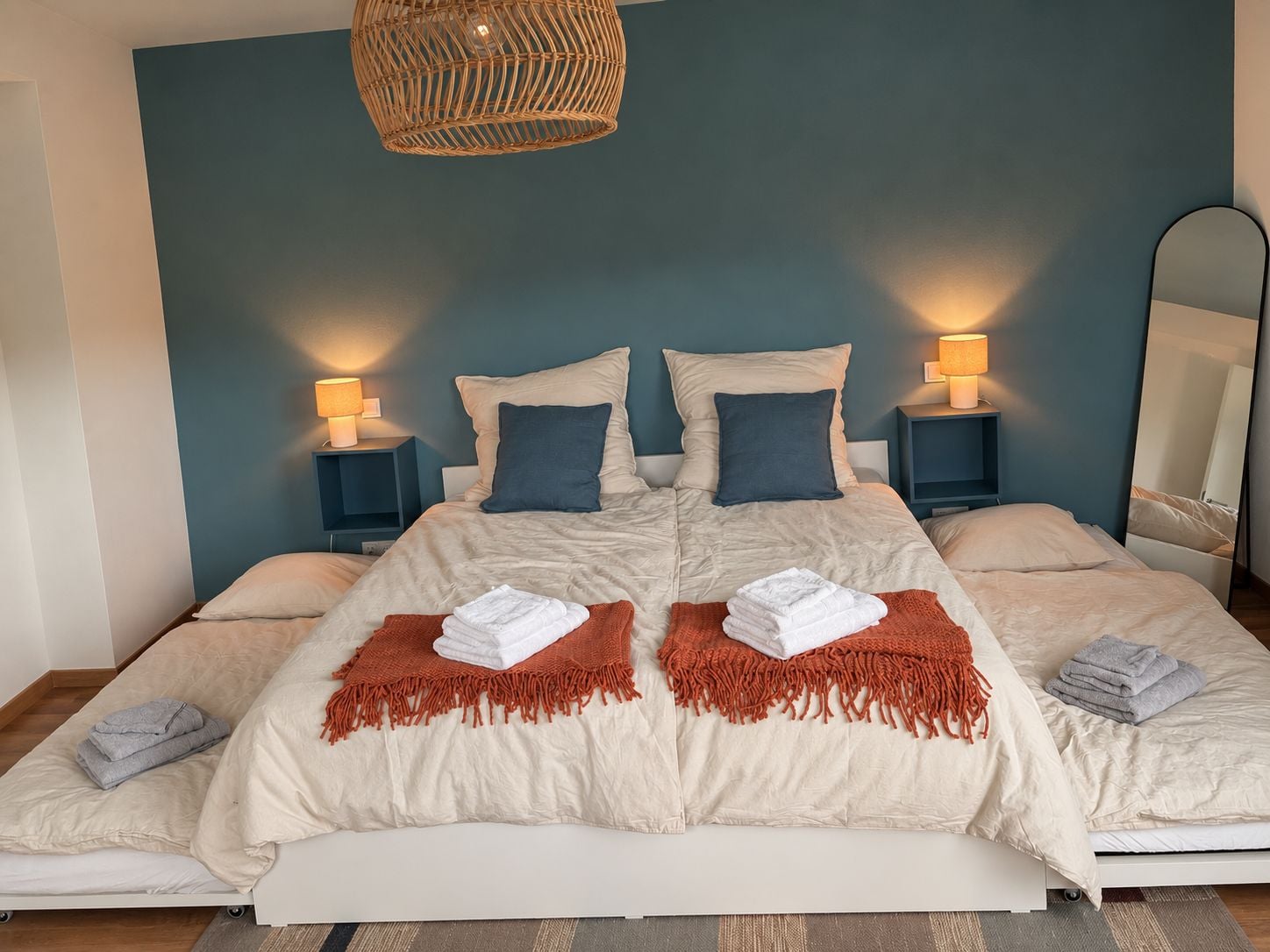 A double bed with a beige and white duvet, two pillows, a woven headboard, bedside lamps, and a mirror.
