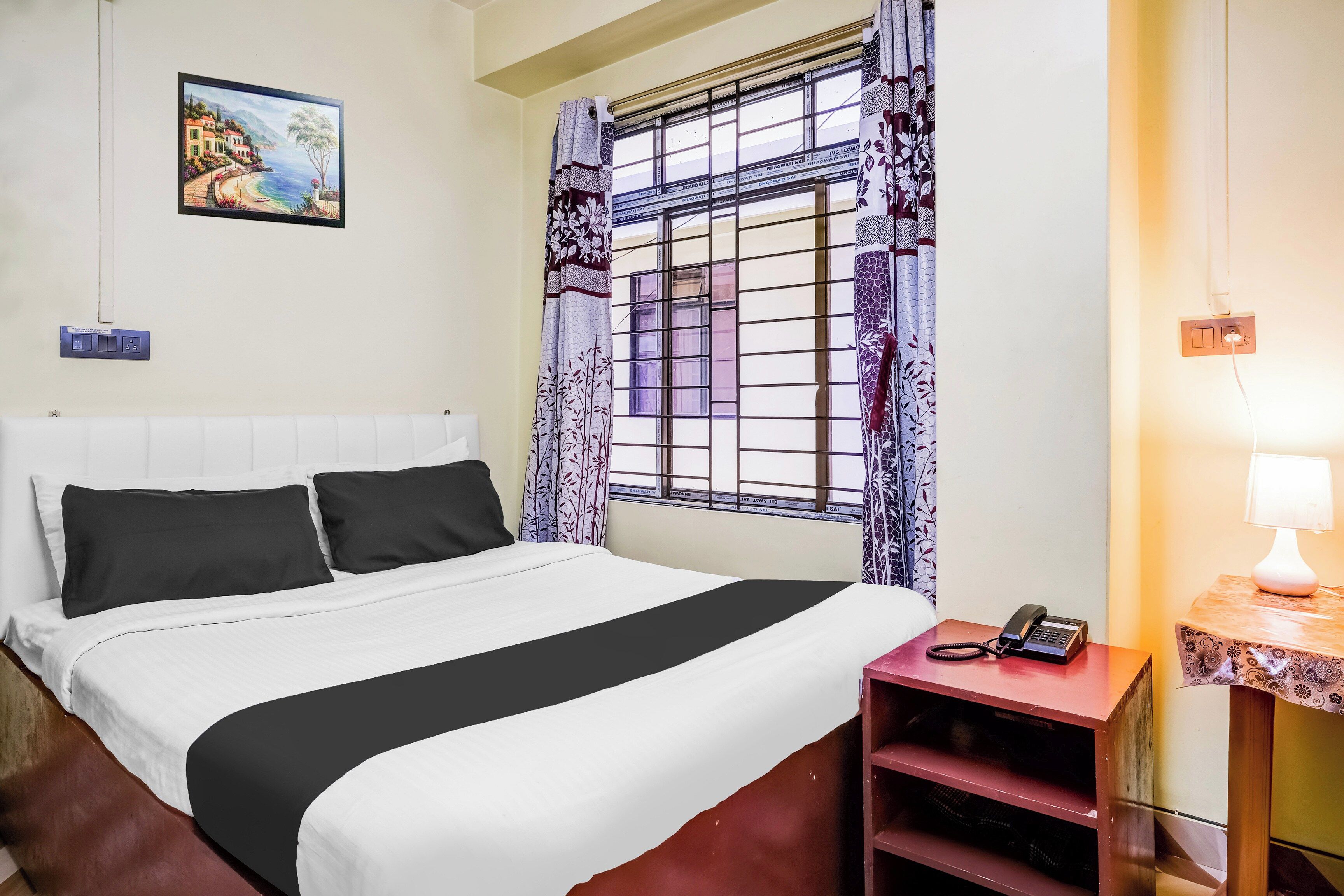 Habitación de hotel con una cama, una mesita de noche con teléfono, una lámpara de pared y una ventana con cortinas estampadas.