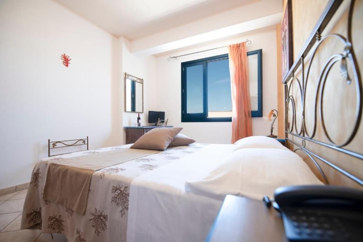 Un dormitorio con cama, mesita de noche, escritorio con silla, ventana con cortinas y un teléfono instalado en la pared.