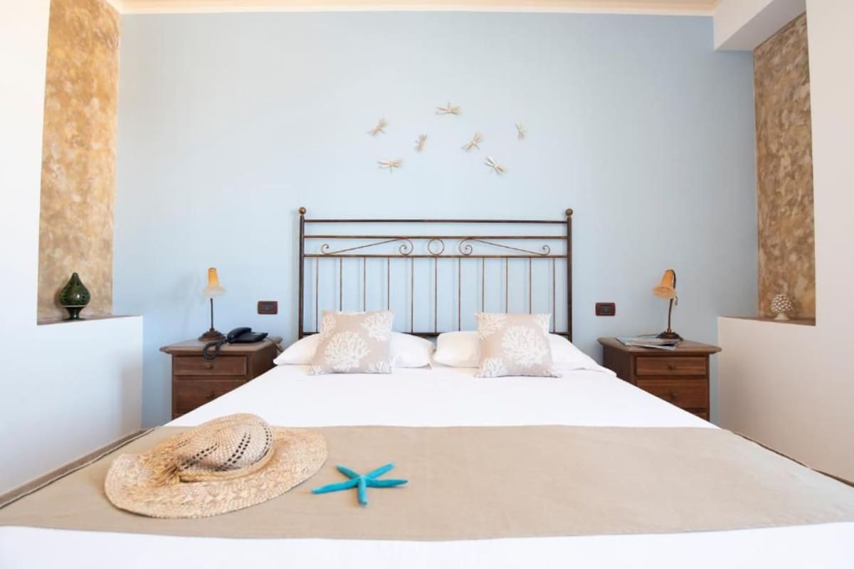 Un dormitorio con cama, dos mesitas de noche, una lámpara de pared, un adorno en la pared y un objeto decorativo con motivos playeros en la cama.