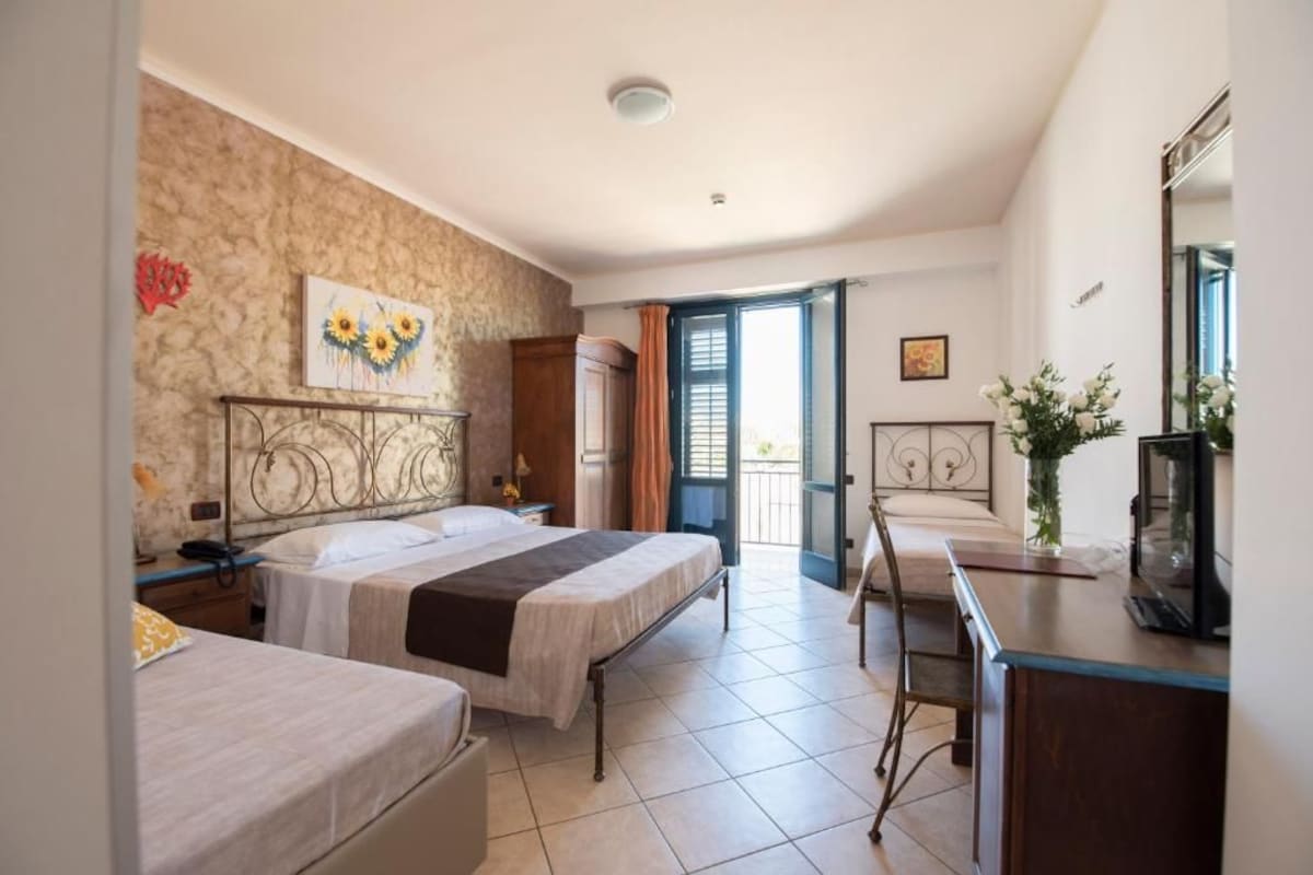 Habitación de hotel con dos camas, un escritorio, una silla, una ventana con cortinas y una puerta que da a un balcón.