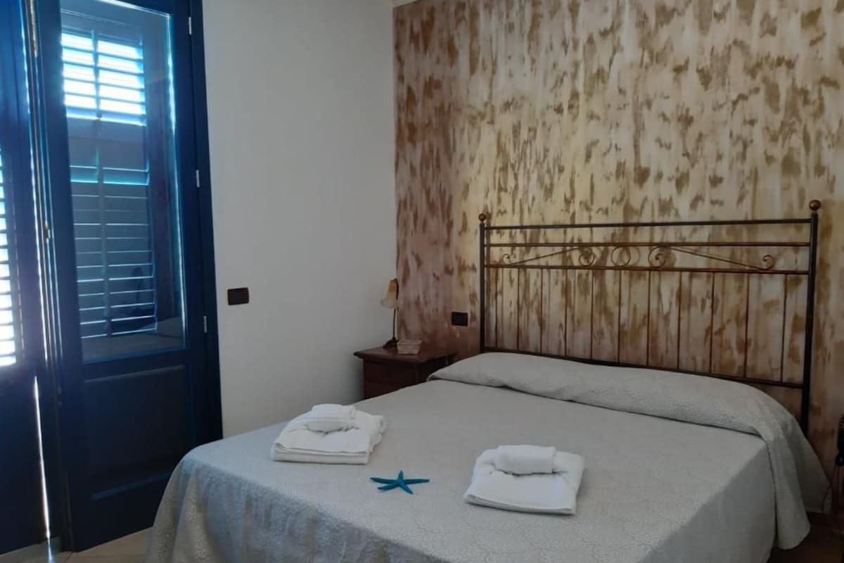 Un dormitorio con cama, mesita de noche, pared con diseño que imita la madera y puerta con persianas.