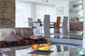 A modern living room with a glass coffee table, a brown sofa, and a dining area in the background. - Holiday apartment with 1 bedroom (Gaienhofen)