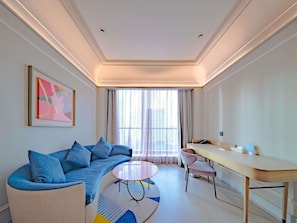 Habitació d'hotel moderna amb un sofà, una taula rodona, un escriptori i una finestra gran amb cortines.