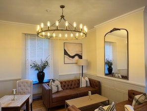 A living room with a brown sofa, a coffee table, a lamp, a chandelier, a mirror, and a painting on the wall. - The Edge Of The Beach (Bude)