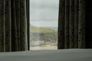 A view through a window with patterned curtains, overlooking a coastal landscape with a boat on the water. - The Edge Of The Beach (Bude)