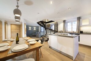 A modern kitchen with a central island, a dining area with a wooden table, and a staircase leading to an upper level. - Heimat1883 - Raftsman's Daughter (Garmisch-Partenkirchen)