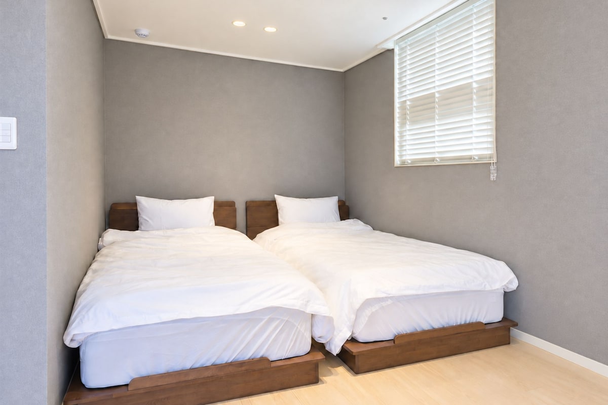 A bedroom with two beds, a window with blinds, and a light switch on the wall.