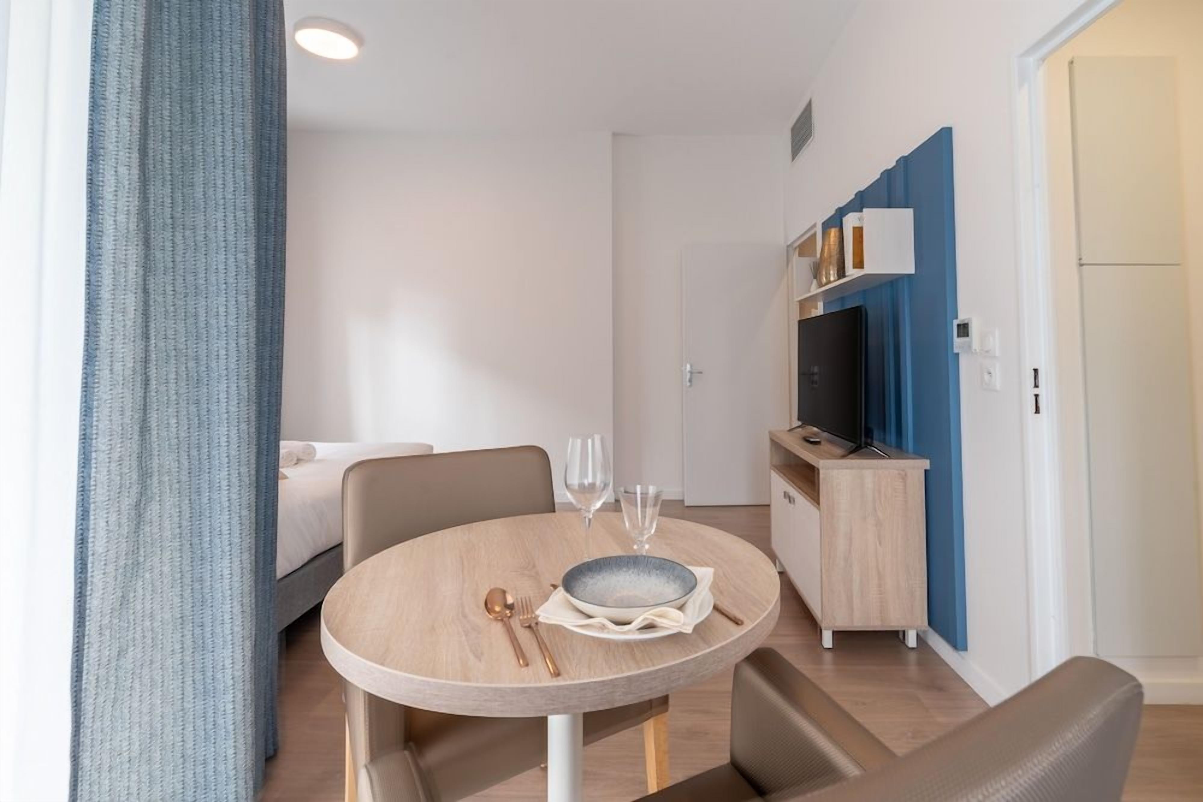 Ein modernes Hotelzimmer mit einem runden Esstisch, einem Bett, einer Küchenzeile und Blick auf die Stadt.