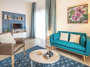 Una sala moderna con un sofá verde azulado, un sillón de cuero marrón, una mesa de centro con una bandeja de objetos, un televisor de pantalla plana y un estante decorativo en la pared.