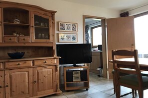 Ein Holzkabinent mit Schubladen, ein Fernseher auf einem Standfuß, ein Esstisch mit einem Stuhl und gerahmte Bilder an der Wand.