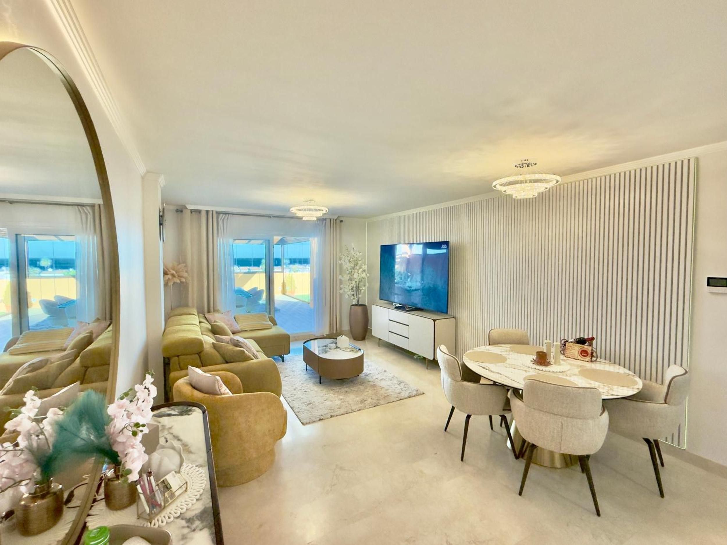 A modern living room with a sofa set, a dining table with chairs, a flat-screen TV, and a large mirror.