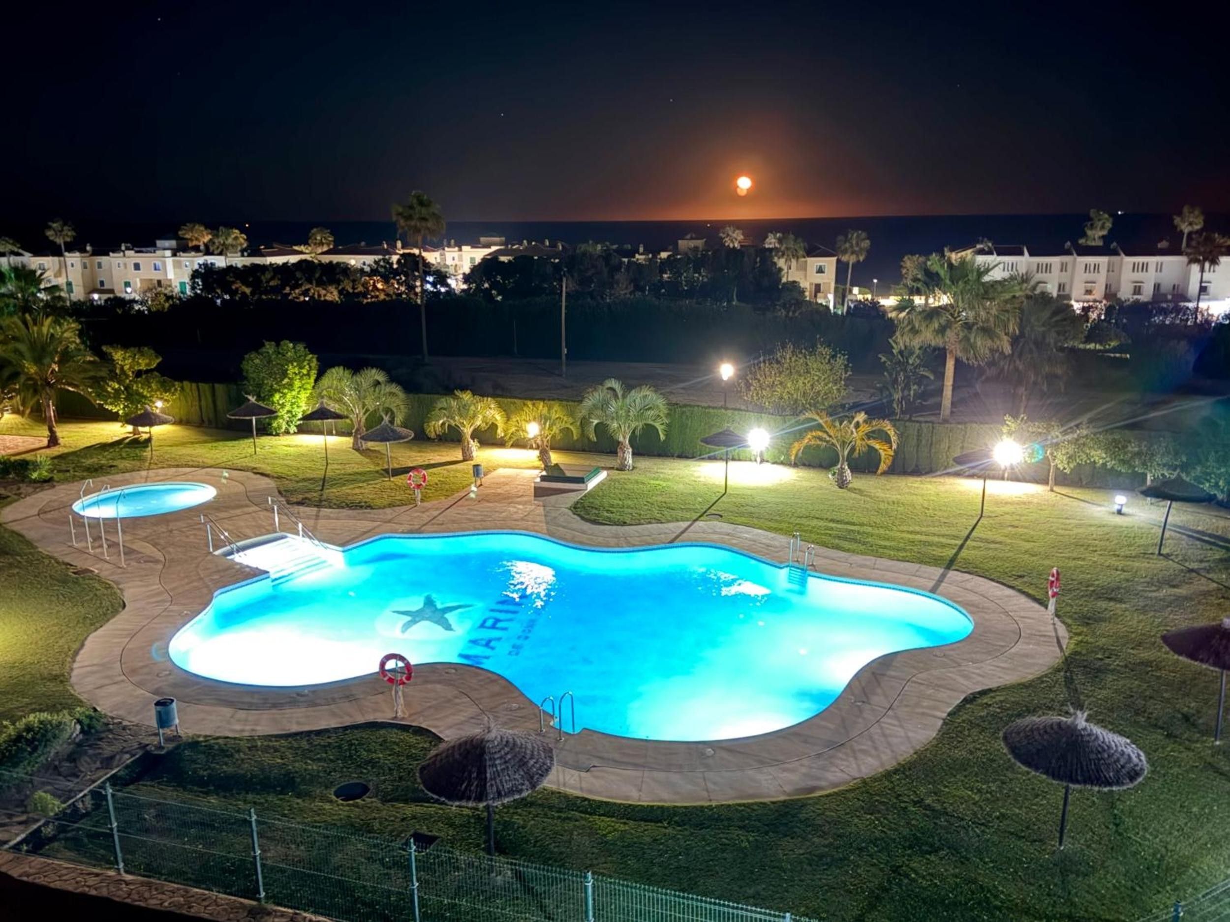 A swimming pool with a star-shaped design, surrounded by a well-lit patio area with umbrellas and a fence.