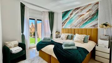 Een moderne slaapkamer met een groot bed, een groene fauteuil, een zacht wit vloerkleed en uitzicht op het groen.