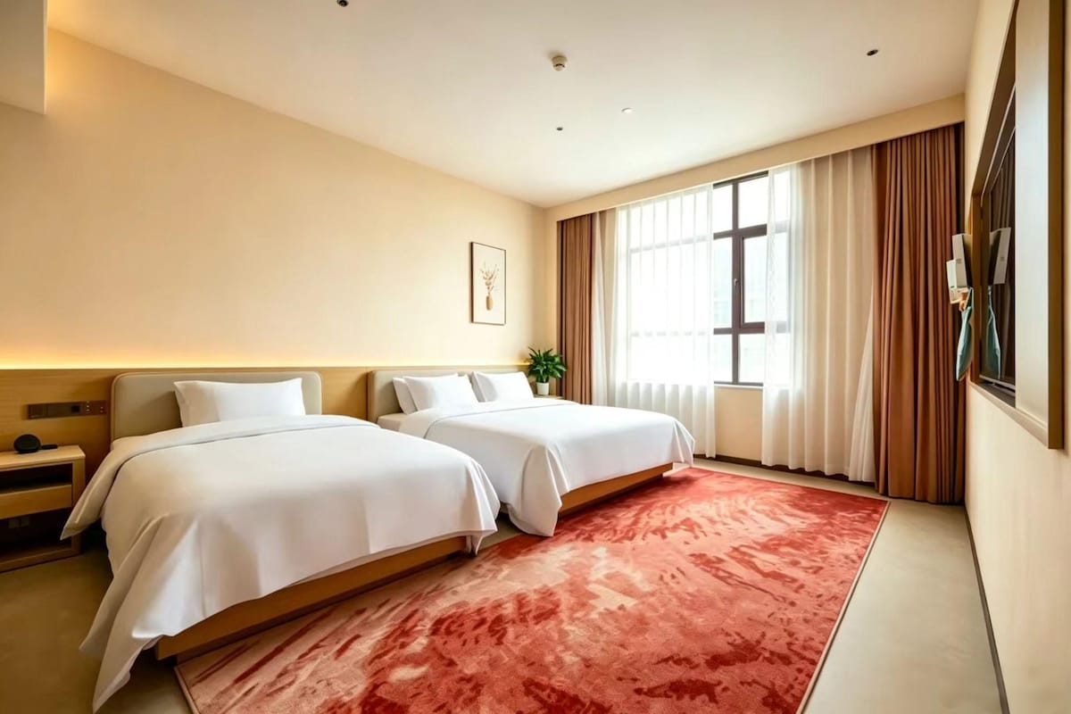 Habitación de hotel con dos camas, un ventanal con cortinas, una alfombra roja y un televisor montado en la pared.