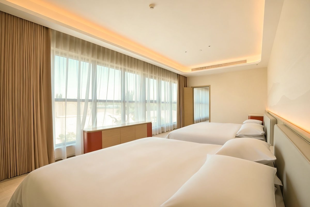 Habitación de hotel con dos camas, un ventanal con cortinas translúcidas y vistas al exterior.