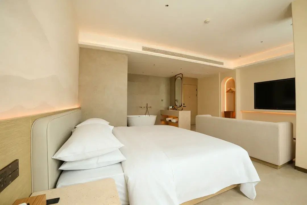 A modern hotel room with a large bed, a seating area, a television, and a bathroom visible in the background.