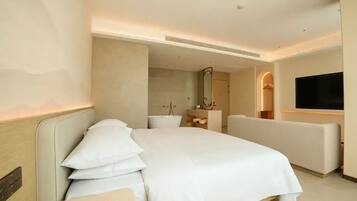 A modern hotel room with a large bed, a seating area, a television, and a bathroom visible in the background.