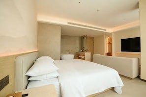 Habitación de hotel moderna con una cama grande, un área de descanso, un televisor y un baño visible al fondo.