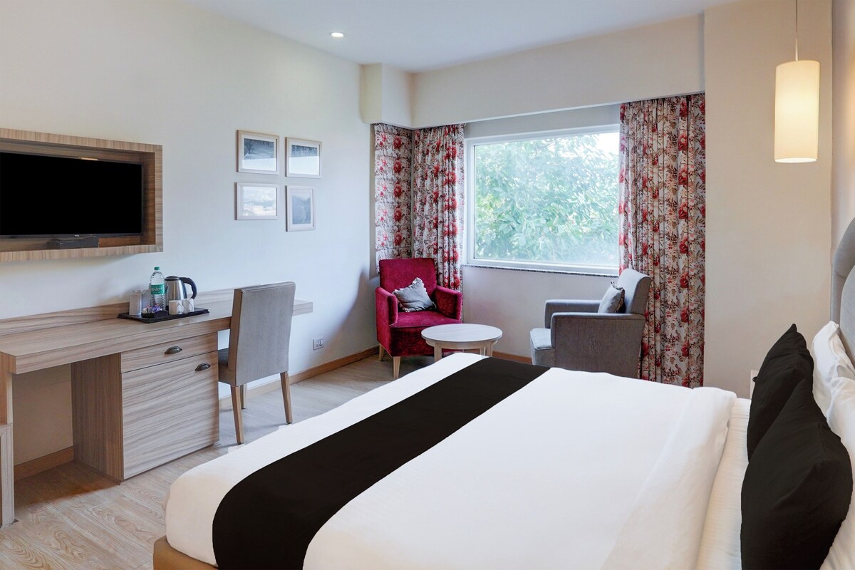Habitación de hotel con cama, escritorio con televisor de pantalla plana, silla, mesita pequeña y ventana con cortinas.