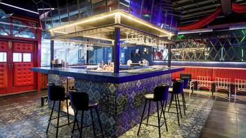 Uma área de bar com balcão revestido de azulejos azuis e roxos, banquetas pretas e uma porta vermelha ao fundo.
