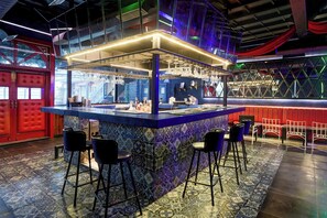 Uma área de bar com balcão revestido de azulejos azuis e roxos, banquetas pretas e uma porta vermelha ao fundo.