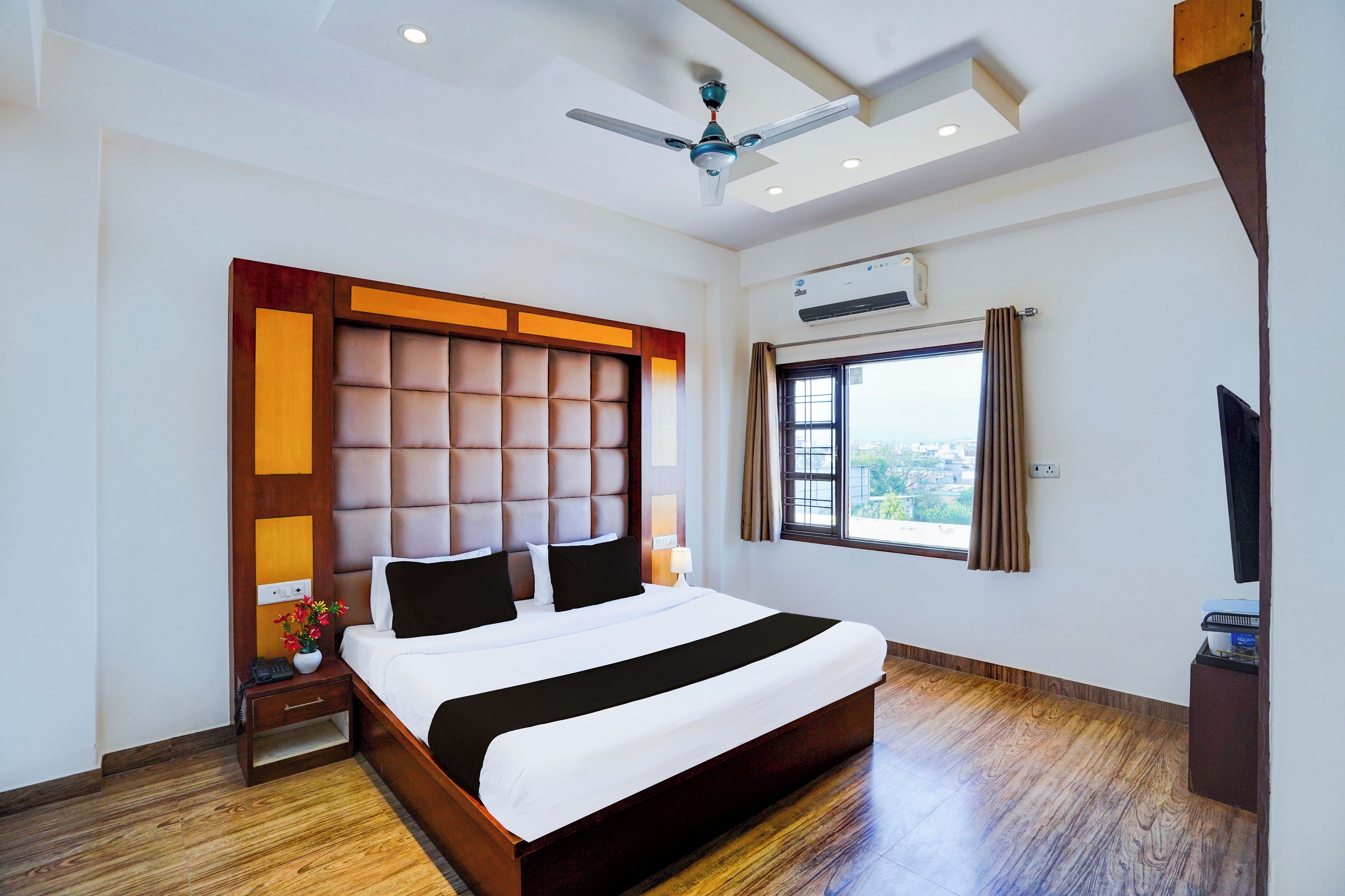 A hotel room with a large bed, a ceiling fan, an air conditioning unit, a window with curtains, and a television.