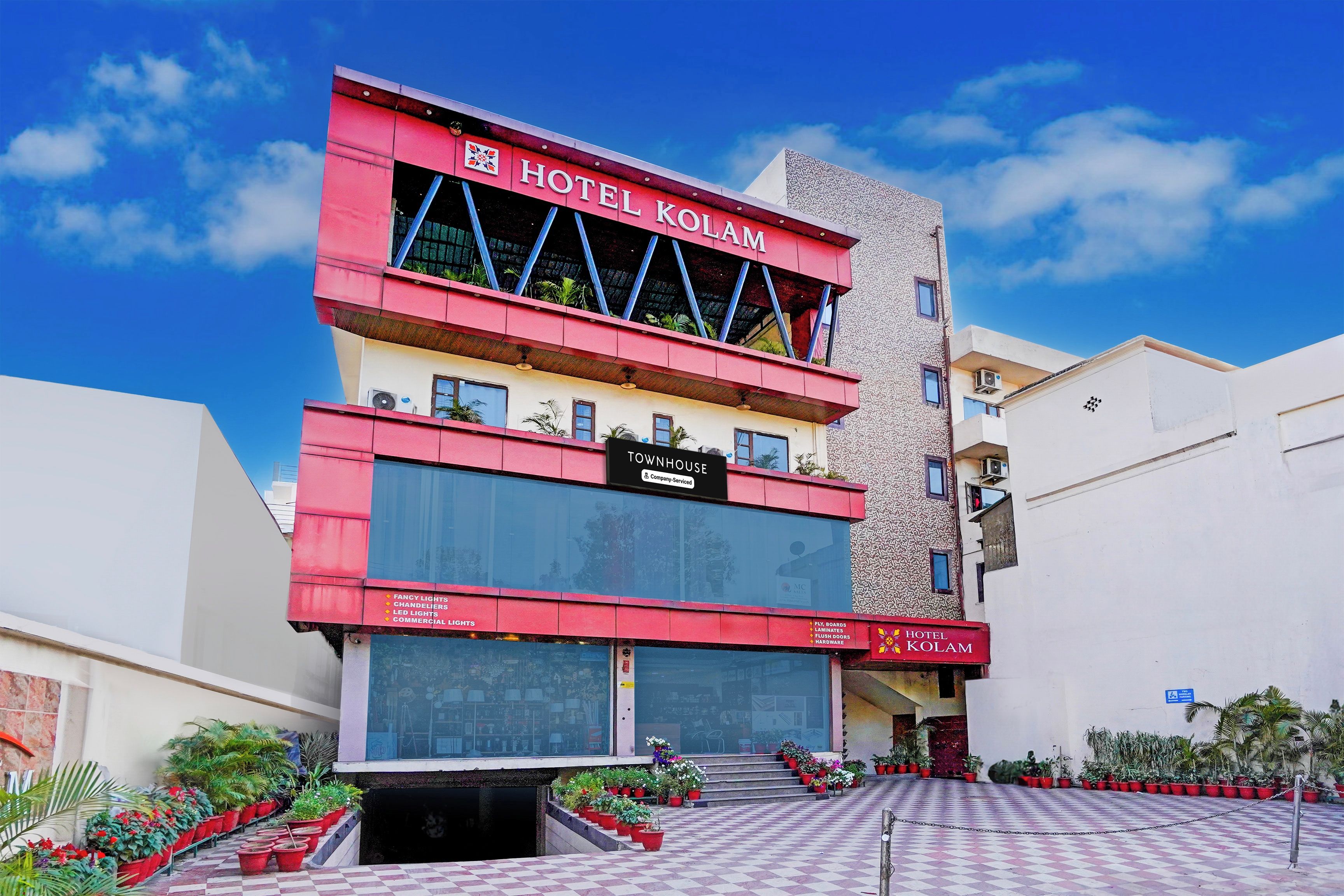 A hotel building with a prominent red and white facade, a sign indicating it's a hotel, and a checkered pavement area with potted plants.