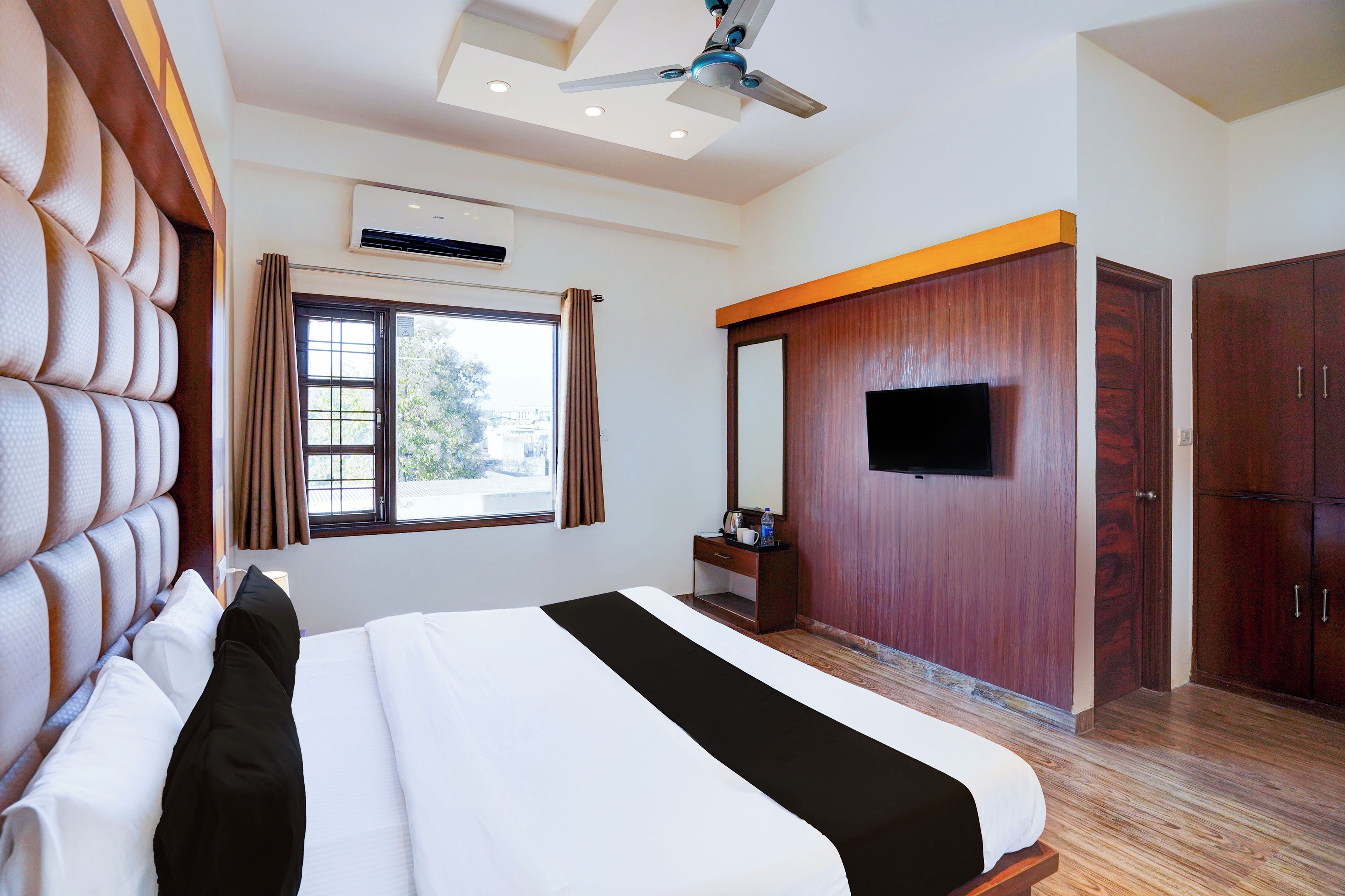 A hotel room with a large bed, a flat-screen TV mounted on a wooden wall, and a ceiling fan.