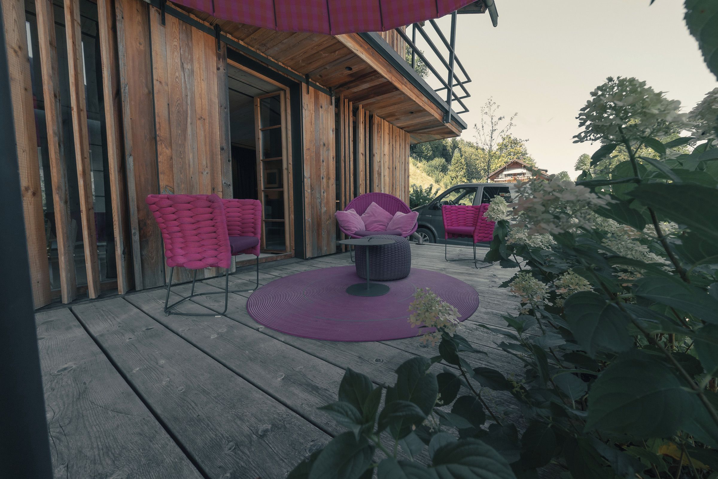 Ein Holzgebäude mit Balkon, zwei rosa Stühlen, einem runden Tisch und einer violetten Matte.
