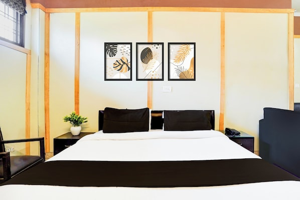 A hotel room with a large bed, two black pillows, a small plant on a nightstand, and three framed artworks on the wall.
