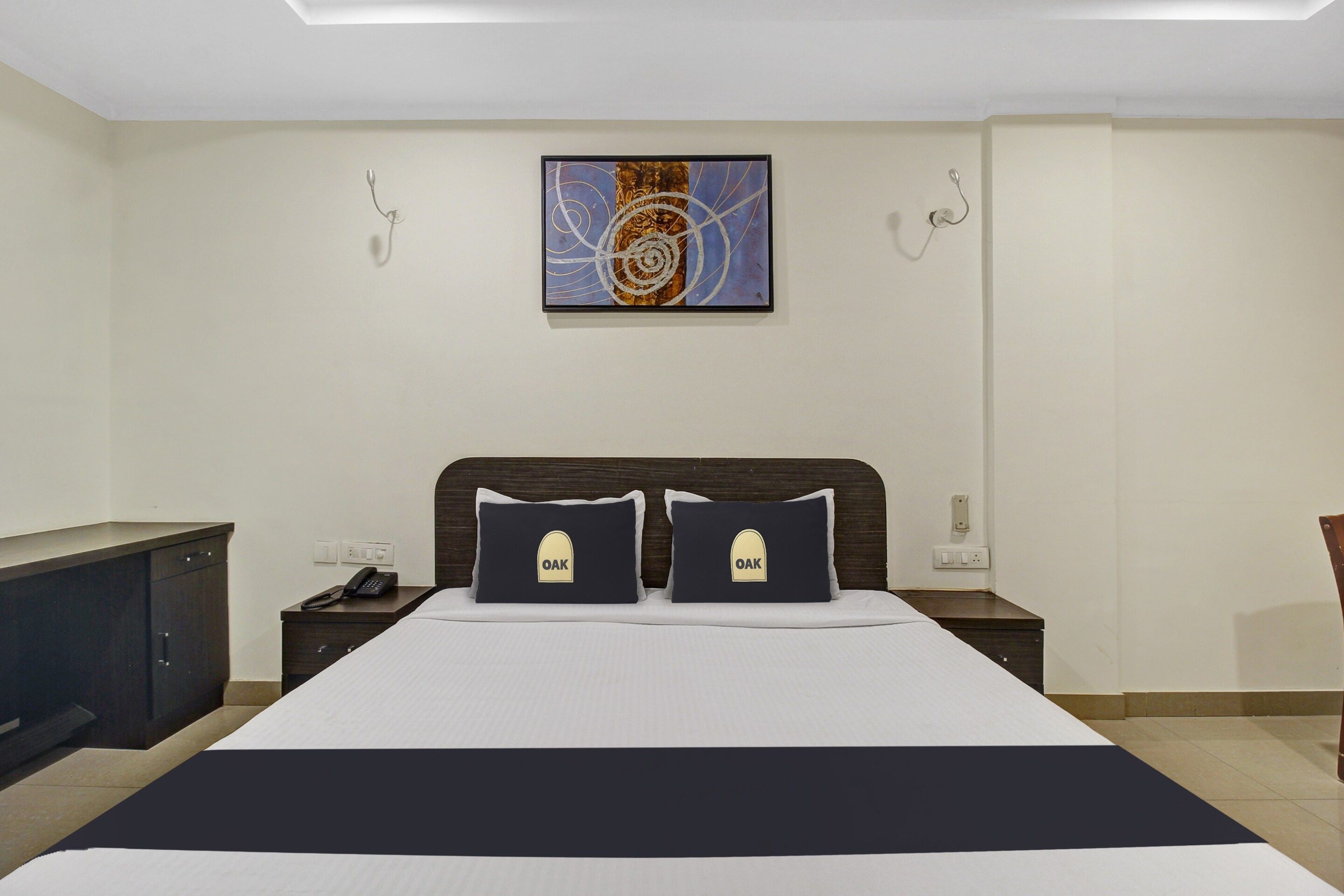 A hotel room with a large bed, two pillows with 'OAK' branding, a bedside table with a phone, and a wall-mounted painting.