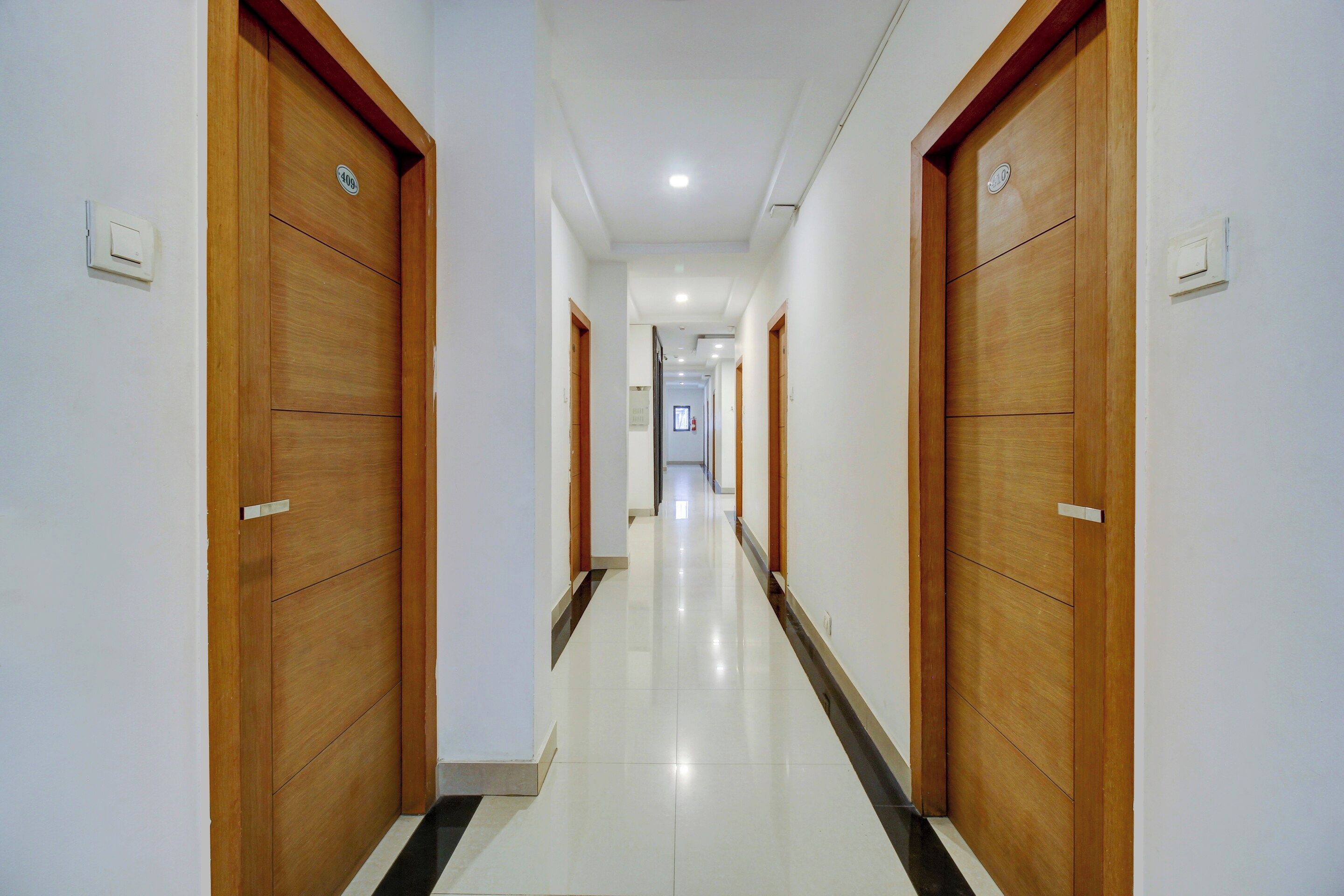 A corridor with wooden doors and a tiled floor, leading to a hallway with a fire extinguisher.