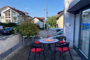 Ett utomhusområde med sittplatser, ett runt bord och stolar, en krukväxt och en byggnad med en synlig entré.