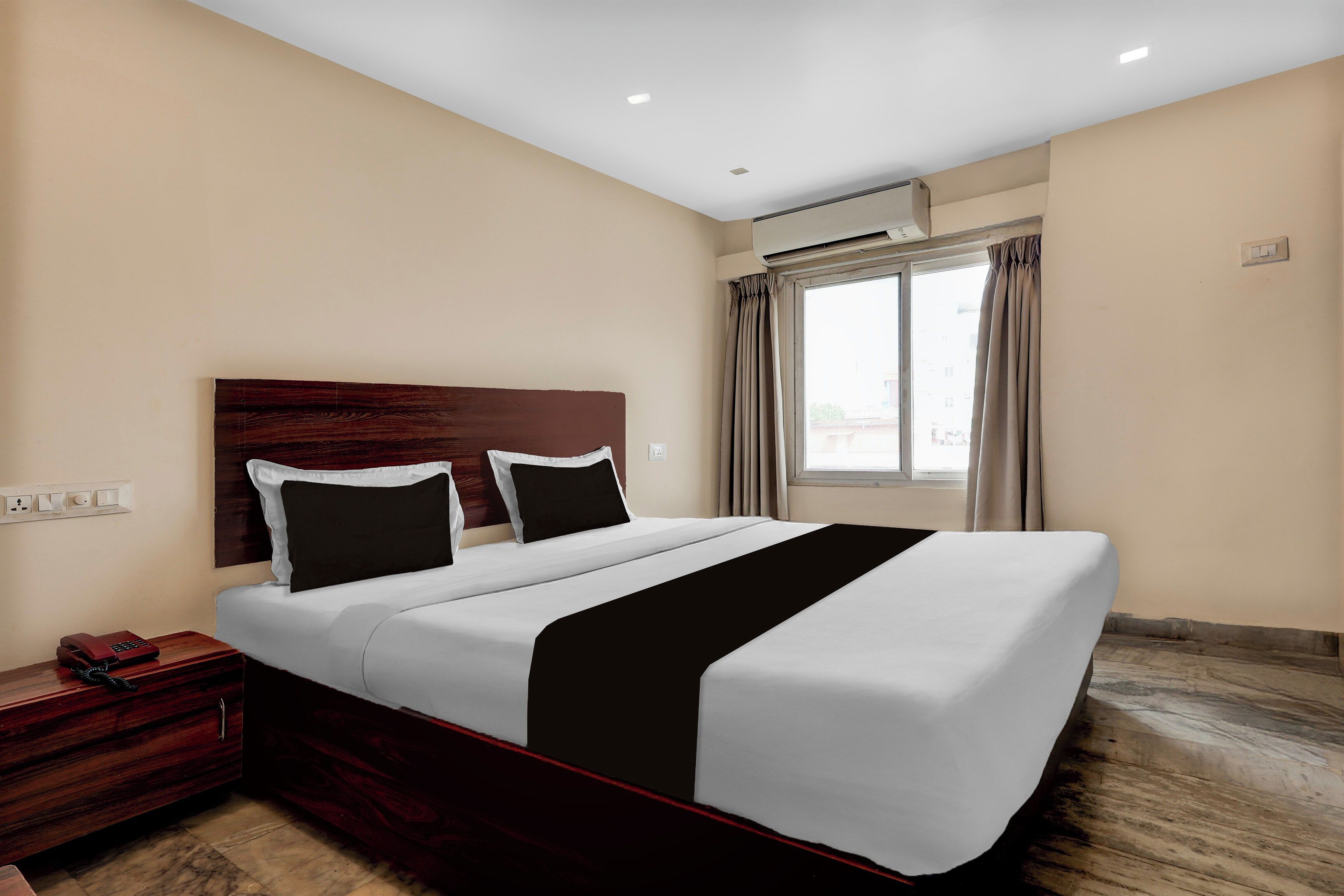 Quarto de hotel com uma cama grande, cabeceira de madeira, mesa de cabeceira com telefone e janela com cortinas.