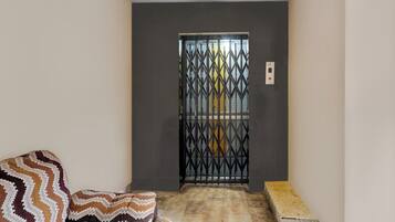 A narrow hallway with a patterned cushion on the left, a metal gate with a yellow panel, and a small bench at the end.