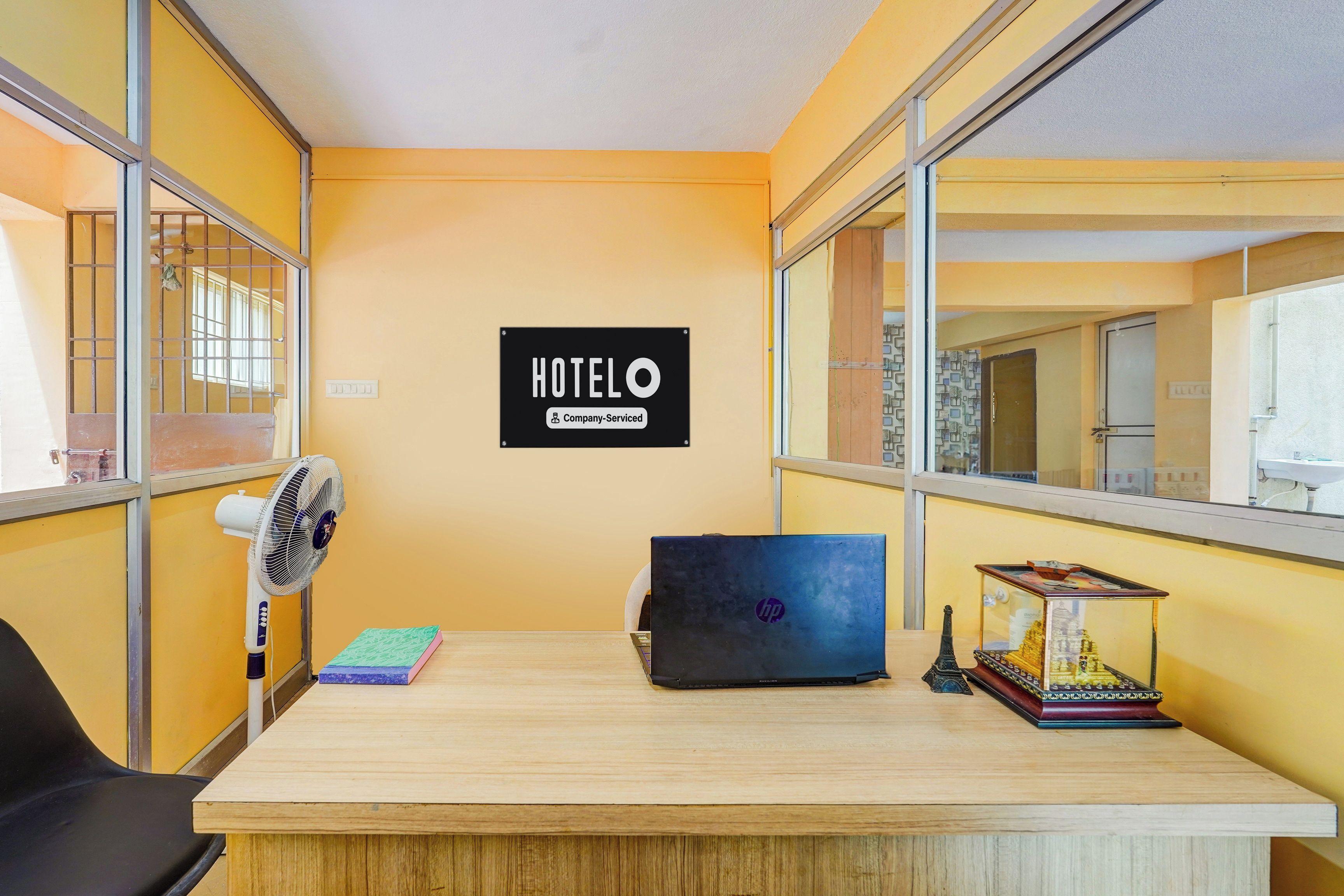 A hotel room with a wooden desk, a laptop, a fan, a small statue, and a sign indicating the room is company-serviced.