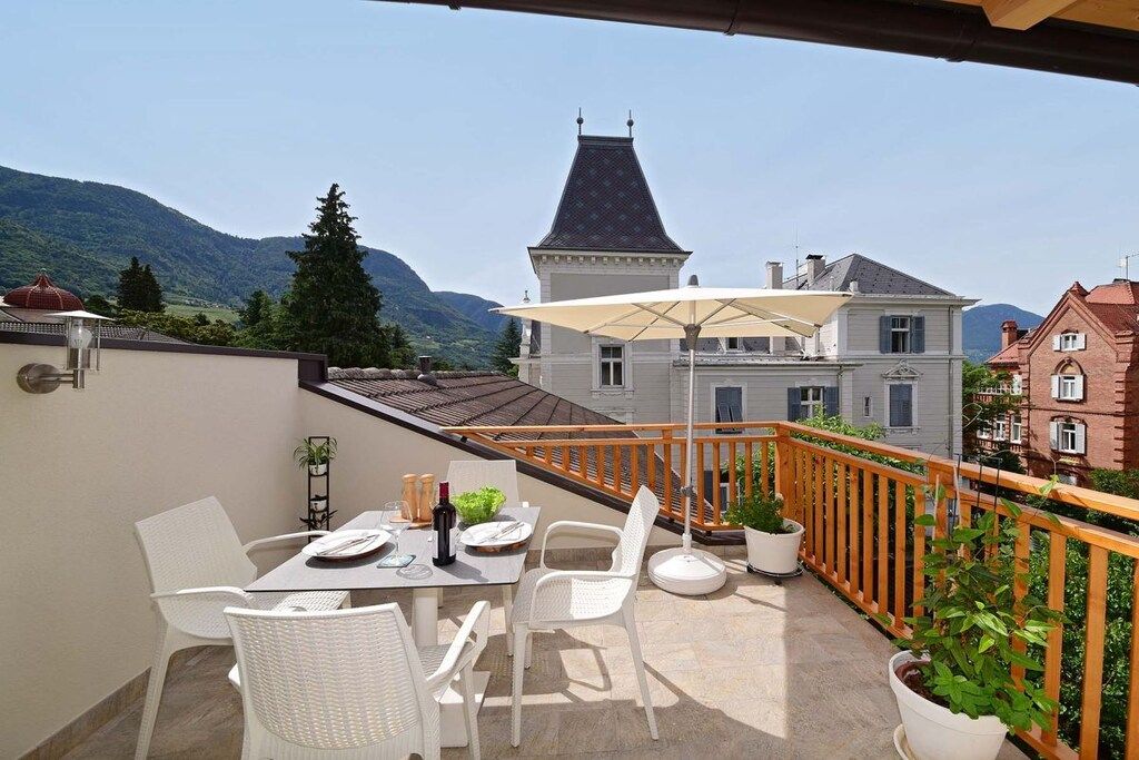 Un balcone con tavolo e sedie, piante in vaso e vista su una montagna e degli edifici.