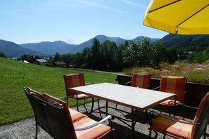 Un área de estar al aire libre con una mesa, sillas, una sombrilla amarilla y un paisaje montañoso de fondo.