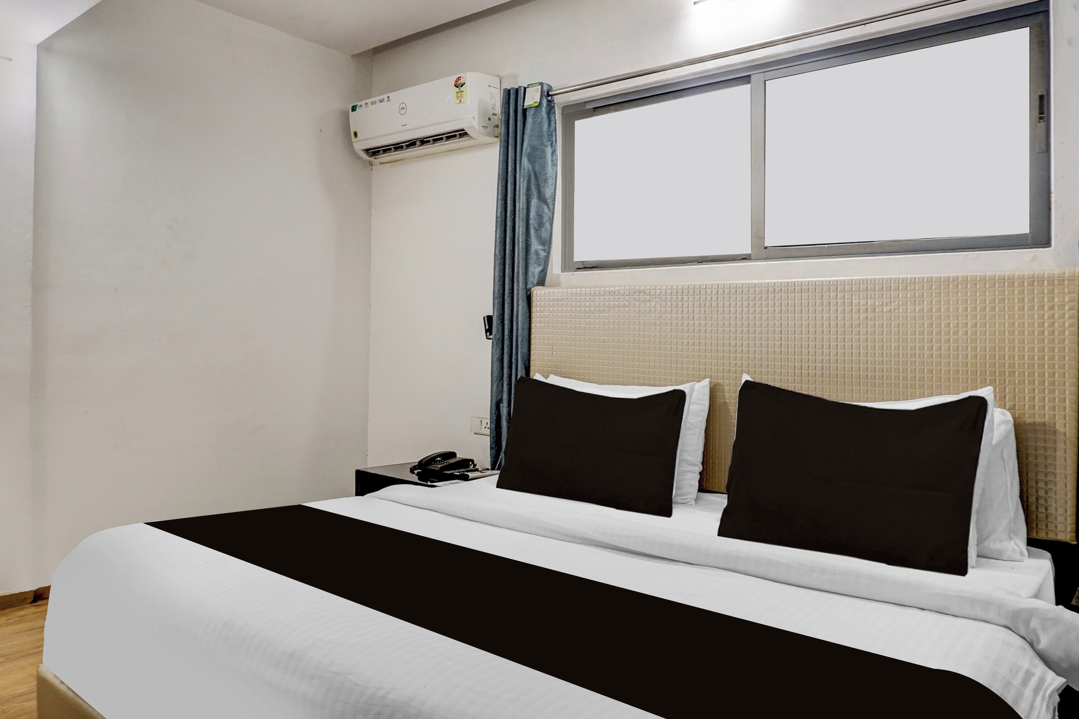 A hotel room with a bed, a bedside table, a telephone, and an air conditioning unit mounted on the wall.