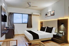 A hotel room with a bed, a desk, a TV, a window with curtains, an air conditioning unit, and artwork on the wall.