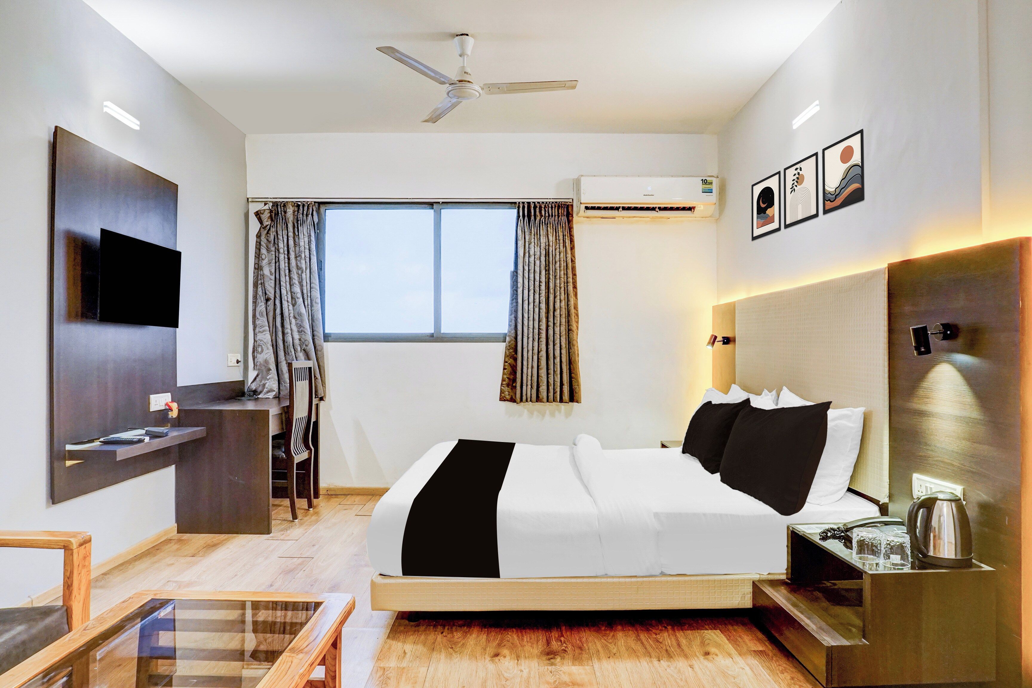 Habitación de hotel con cama, escritorio, silla, televisor, ventilador de techo, aire acondicionado y ventana con cortinas.