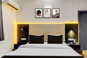 A hotel room with a large bed, two bedside tables, an air conditioning unit, and three framed artworks on the wall.