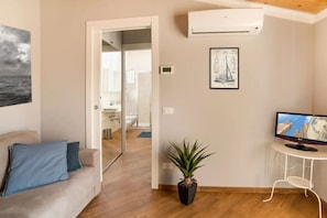 A living room with a sofa, a potted plant, an air conditioner, a framed picture, and a television. - In the heart of the city, adjacent to the central Piazza del Popolo, at the gates of the historic center and close to the tree-lined avenue that leads to the sea, Il Maestrale is a delightful newly built attic, ready to accommodate up to 4 people.Withi (Albenga)