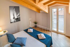 A bedroom with a bed, pillows, a blue towel, a sailboat painting, a small plant, and a balcony with a door. - In the heart of the city, adjacent to the central Piazza del Popolo, at the gates of the historic center and close to the tree-lined avenue that leads to the sea, Il Maestrale is a delightful newly built attic, ready to accommodate up to 4 people.Withi (Albenga)