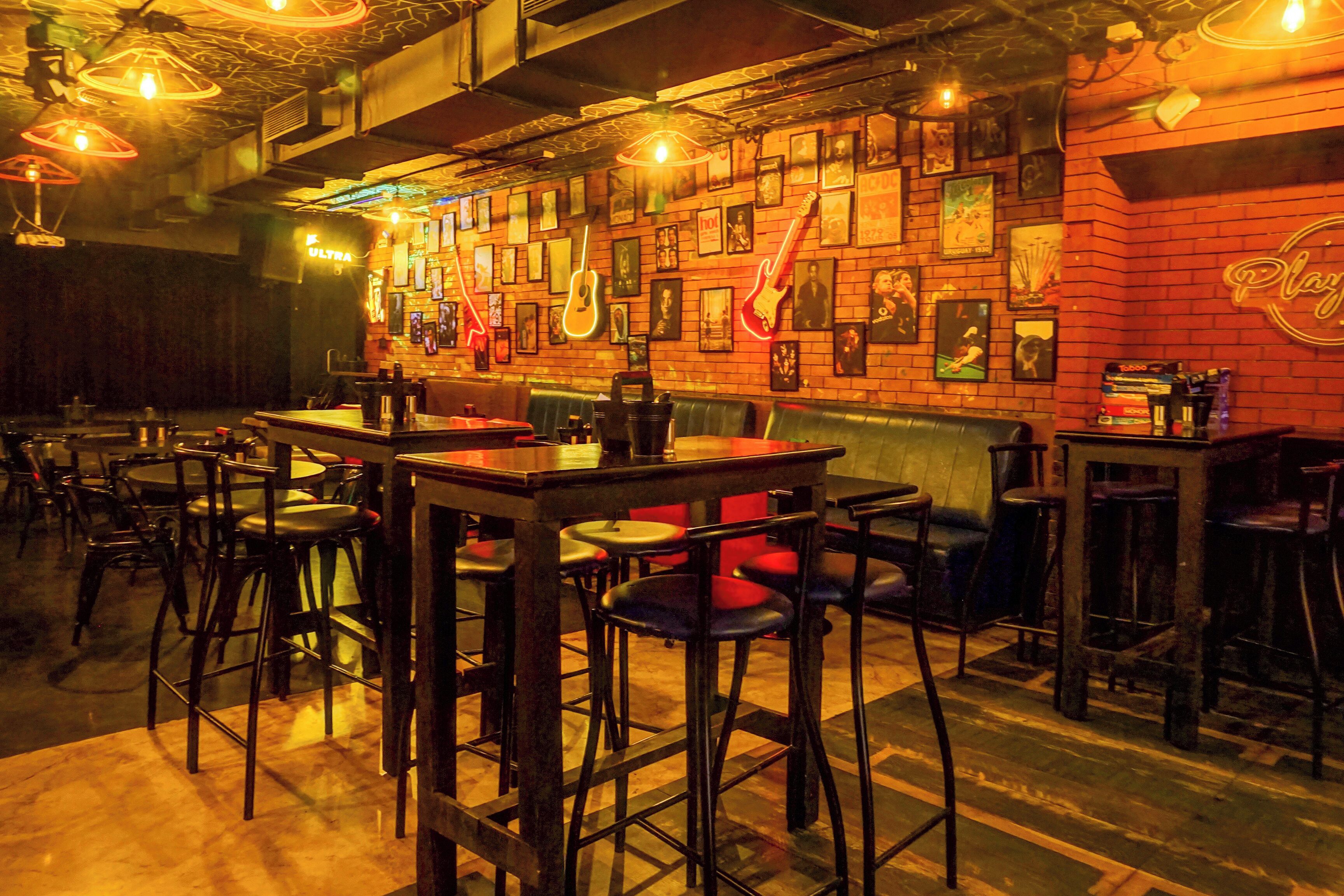 A bar area with multiple stools and tables, a brick wall with framed pictures, and a neon sign.